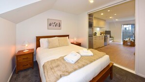 A bedroom with a bed, bedside tables, a nightstand, a wall-mounted picture, and an adjoining kitchen area. - City Fringe Stay with Easy CBD Access (Christchurch)