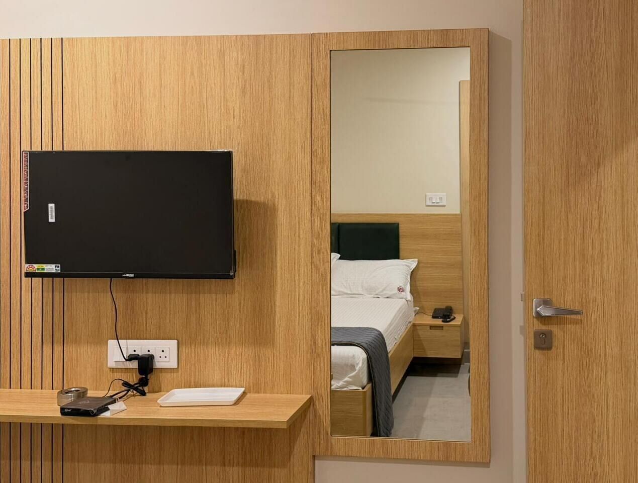 A hotel room with a wooden panel wall, a flat-screen TV mounted above a wooden desk, a bed with a green headboard, and a mirror reflecting the room.