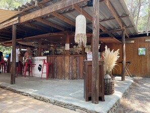 Un espacio de bar al aire libre de estilo rústico, con vigas de madera, una barra y taburetes rojos.