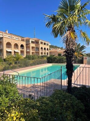 A pool area with a palm tree, a building with arched balconies, and a clear blue sky. - T2 sea and mountain view, large terrace ☀️ with swimming pool - 5 mn des Plages 🏖️ (L'Île-Rousse)