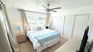Un dormitorio con cama, ventilador de techo, mesita de noche con lámpara, ventana con cortinas y armario.