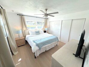 Ein Schlafzimmer mit Bett, Deckenventilator, Nachttisch mit Lampe, Fenster mit Vorhängen und einem Kleiderschrank.