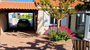 A covered driveway leading to a house with a parked car, a small garden with pink flowers, and a striped chair. - La Pause Jardaise near beaches (Jard-sur-Mer)