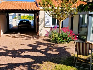 Ein überdachter Weg führt zu einem Haus mit einem parkenden Auto, einem kleinen Garten mit rosa Blumen und einem gestreiften Stuhl.