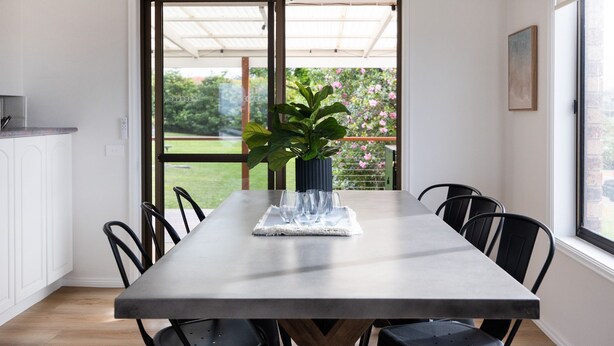 Un comedor con una mesa, sillas, una planta en maceta y vistas de un jardín a través de un ventanal.