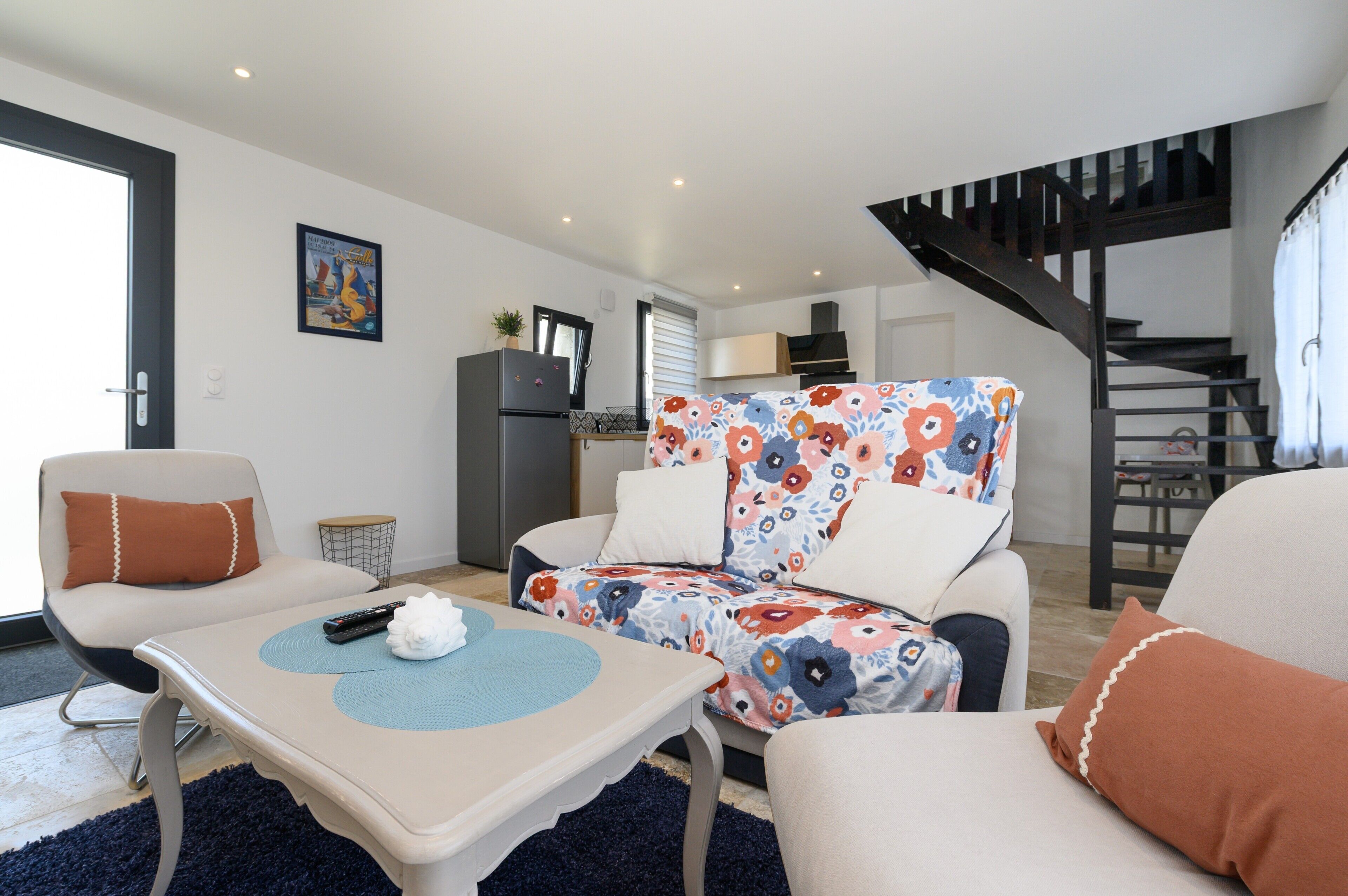 Un salon comprenant un canapé, un fauteuil et une table basse, un coin cuisine avec un réfrigérateur, et un escalier.