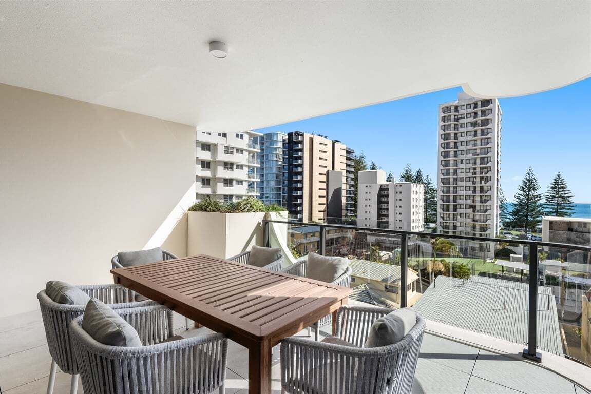 A balcony with a wooden table and wicker chairs, overlooking a cityscape with tall buildings and a clear sky.