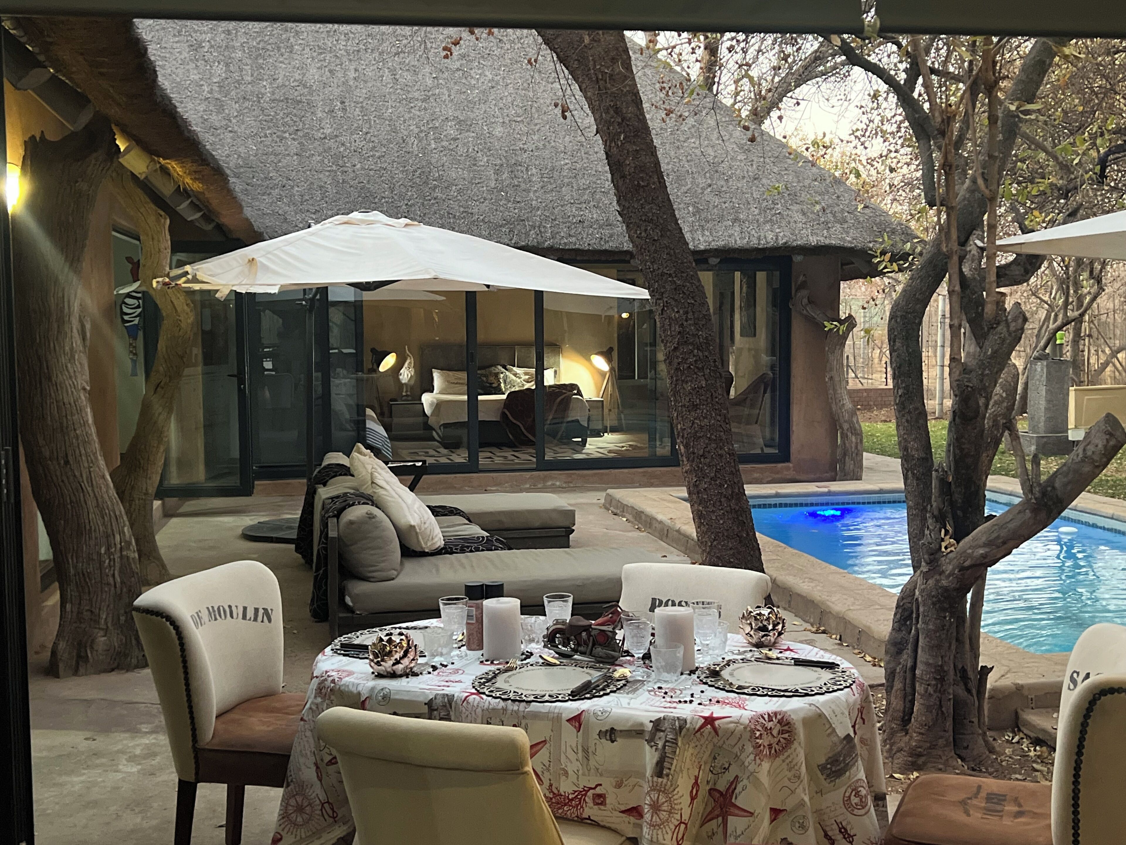Un'area pranzo all'aperto con un tavolo apparecchiato per due, circondata da sedie, una piscina e una stanza con letto interamente in vetro.