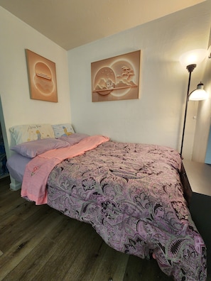 Ein Schlafzimmer mit einem Bett, Kissen, einer Decke, einer Wandlampe und zwei gerahmten Kunstwerken.