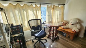 Una habitación con suelo de madera, una silla de oficina negra, una mesa de madera, un oso de peluche y un ventanal con cortinas.