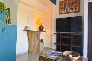 Ein Wohnzimmer mit Fernseher, Lampe, Couchtisch mit Zeitung, Pflanze und einer Wandregal.