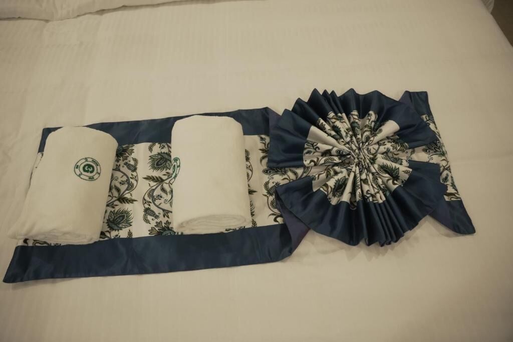 Una cama ordenada con una colcha azul y blanca estampada, dos toallas enrolladas y una almohada decorativa azul y blanca.