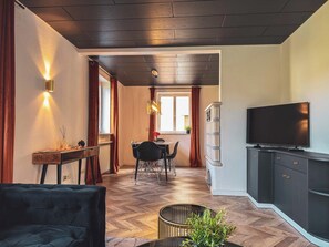 Ein modernes Wohnzimmer mit dunklem Holzboden, einem schwarzen Sofa, einem Essbereich mit Tisch und Stühlen sowie einem an der Wand montierten Fernseher.