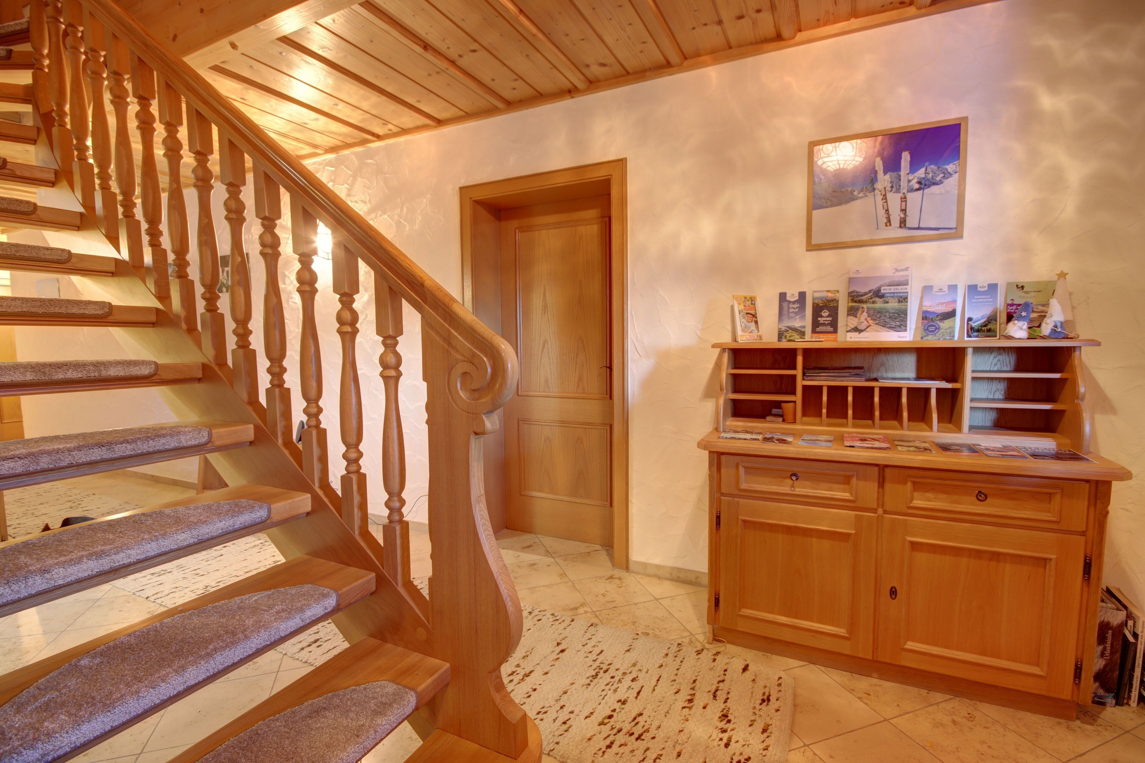 A wooden staircase with a handrail leading to a door. A wooden cabinet with a shelf displaying books and a framed picture hangs on the wall.