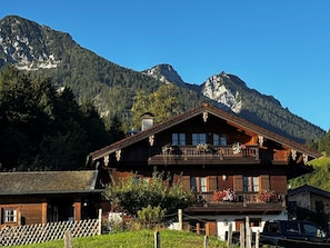 A traditional wooden chalet with a balcony, surrounded by a wooden fence and a mountain backdrop.