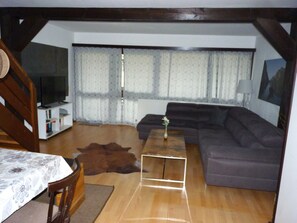 Una sala de estar con sofá, televisor, mesa de madera y un ventanal con cortinas.