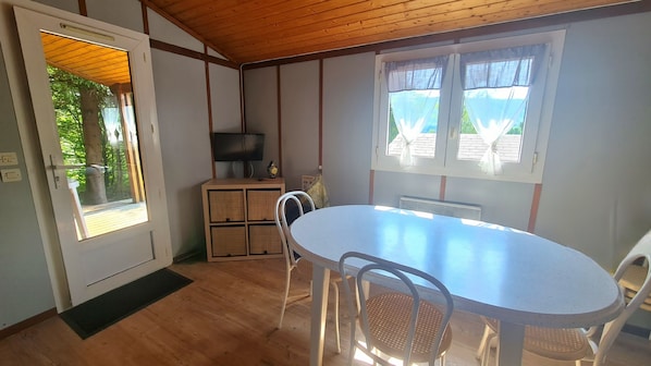 Une pièce avec un plafond en bois, une table blanche avec quatre chaises, un téléviseur posé sur un meuble et une fenêtre avec des rideaux.