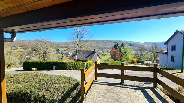 Un espacio al aire libre cubierto con una cerca de madera, un sendero pavimentado y vistas a un pueblo con casas y colinas al fondo.