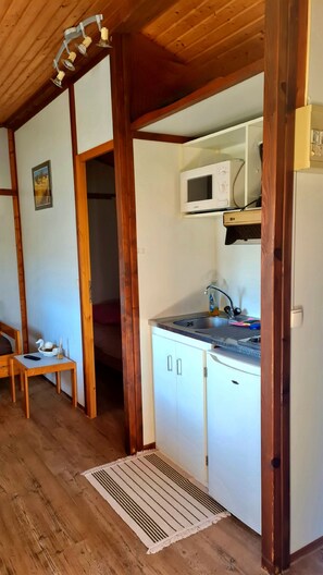 Cocina con microondas, lavamanos y armarios. Se ve una mesa de madera con un control remoto y una cesta.
