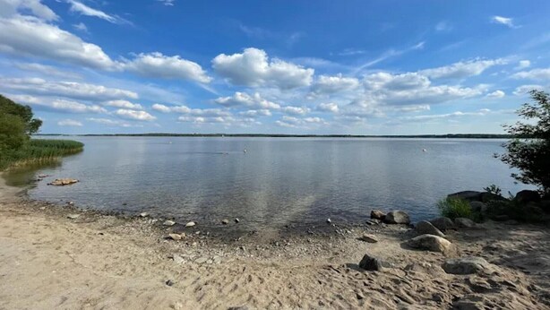 Ein ruhiger See mit sandigem Ufer, umgeben von Grünflächen und Felsen.