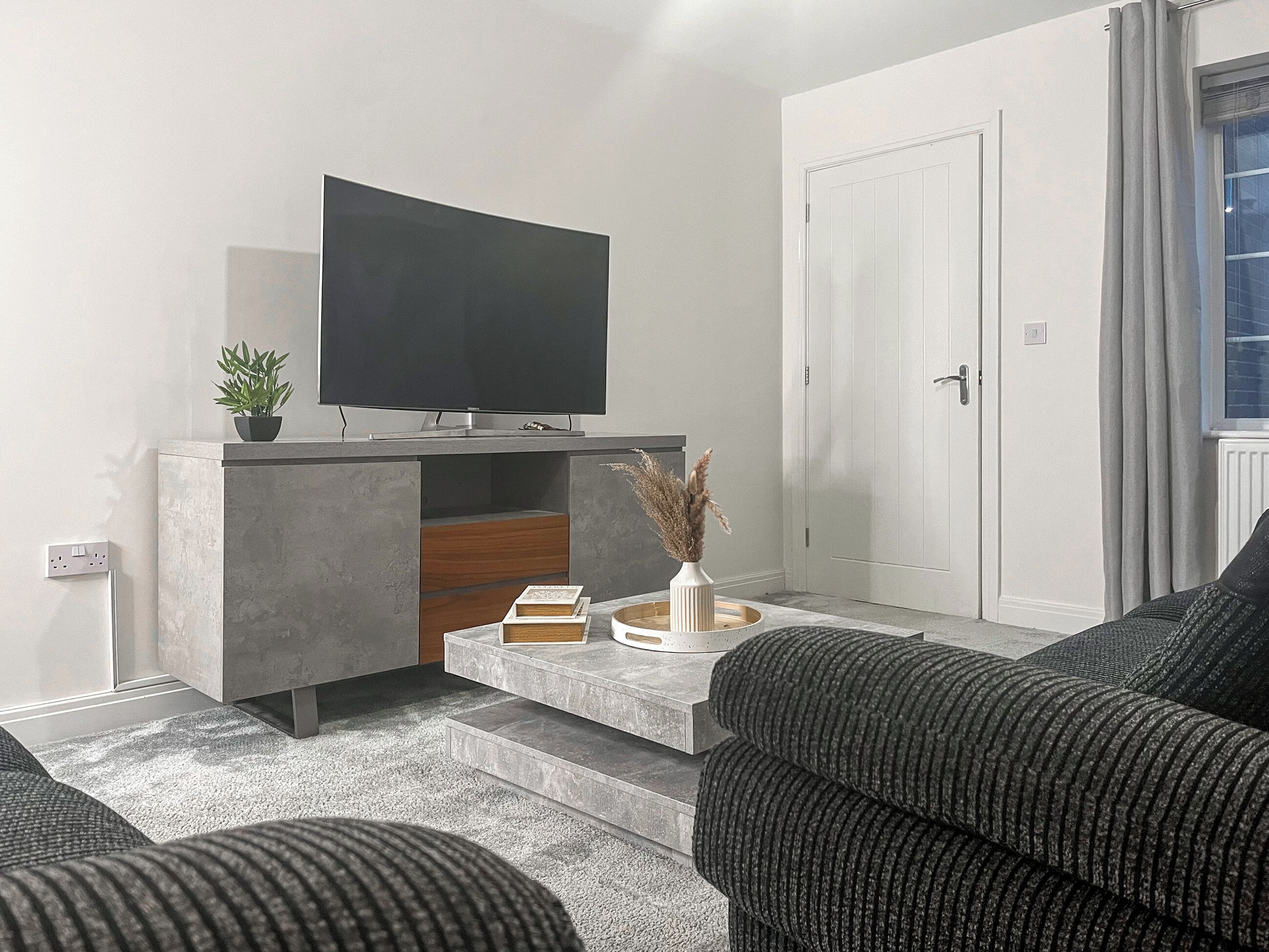 A modern living room with a flat-screen TV, a coffee table with books and a vase, a sofa, and a door.