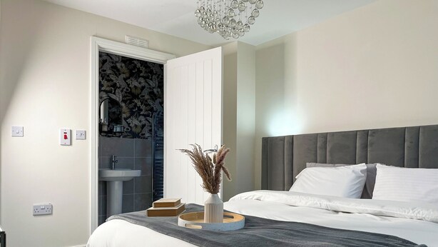 Ein modernes Schlafzimmer mit grauem gepolsterten Kopfteil, einem großen Bett mit weißer Bettwäsche, einem Nachttisch mit Büchern und einer Vase sowie einem Badezimmer mit Waschbecken und Spiegel.