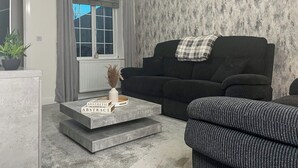 Ein Wohnzimmer mit einer Couch, einem Couchtisch mit Büchern, einer Topfpflanze und einem Fenster mit Vorhängen.