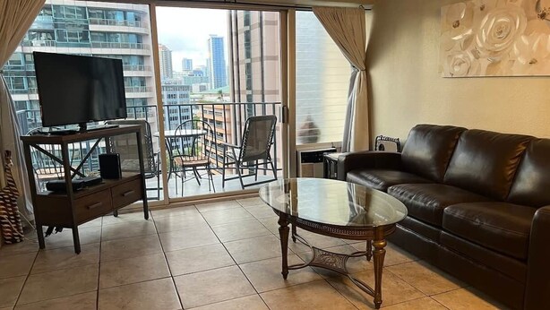 Ein Wohnzimmer mit Glastisch, Ledersofa, Fernseher auf einem Schrank und einem Balkon mit Blick auf die Stadt.