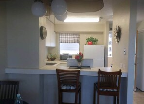 A compact kitchen with a white countertop, two wooden chairs, a microwave, a refrigerator, and a window with a checkered curtain. - Waikiki condo/2 bedroom/FREE parking /4beds (Honolulu)