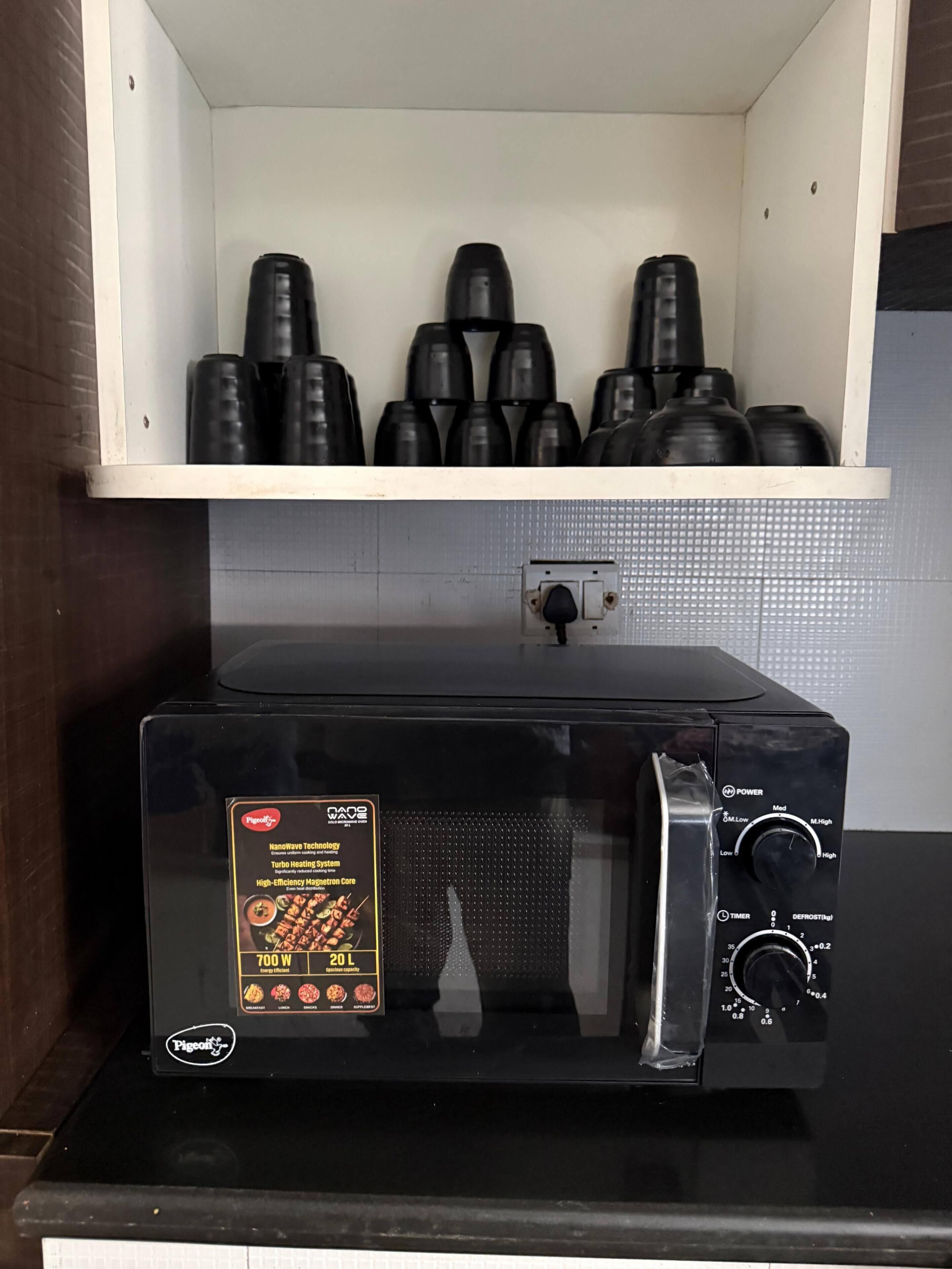 Une cuisine avec un four à micro-ondes posé sur un plan de travail noir, une étagère blanche au-dessus, sur laquelle sont rangés des récipients noirs, et une prise électrique au mur.
