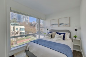 A bedroom with a large window, a bed with a blue and white bedspread, two wall-mounted artworks, and a bedside table with a lamp.