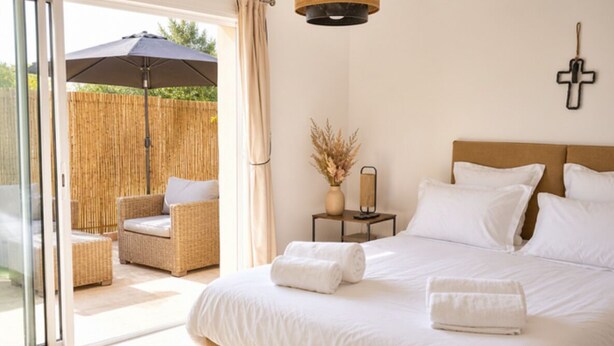 Une chambre à coucher avec un lit, deux oreillers, une croix au mur, un vase contenant des plantes séchées et une chaise en osier à l’extérieur.
