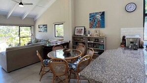 A spacious living area with a dining table, chairs, a sofa, a ceiling fan, a hanging light fixture, a bookshelf, and a clock on the wall.