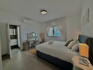 A modern hotel room with a large bed, bedside tables, a desk with a chair, a mirror, and a window with curtains. - Luxury Beach Front Flat Kyma 3 (Germasogeia)