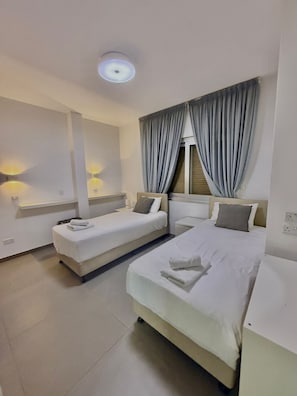 A hotel room with two beds, a large window with curtains, wall-mounted lights, and a ceiling light. - Luxury Beach Front Flat Kyma 3 (Germasogeia)