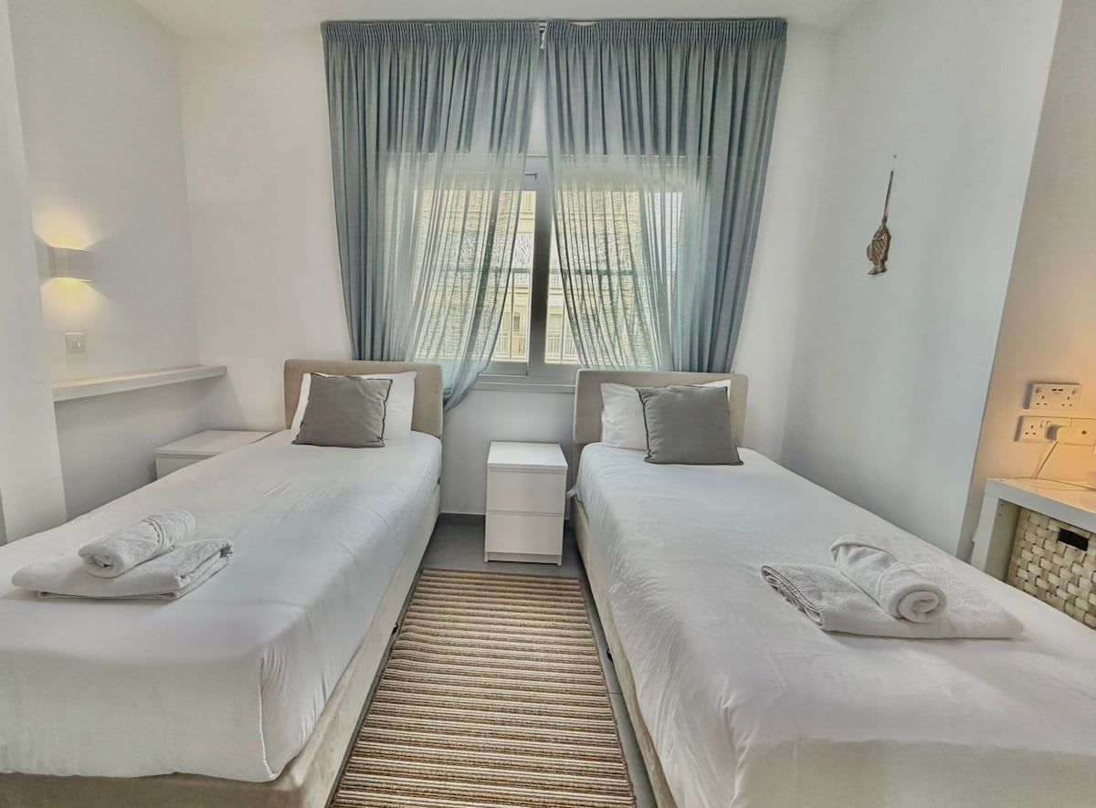 Een hotelkamer met twee enkele bedden, een klein nachtkastje, een raam met gordijnen en een wandlamp.
