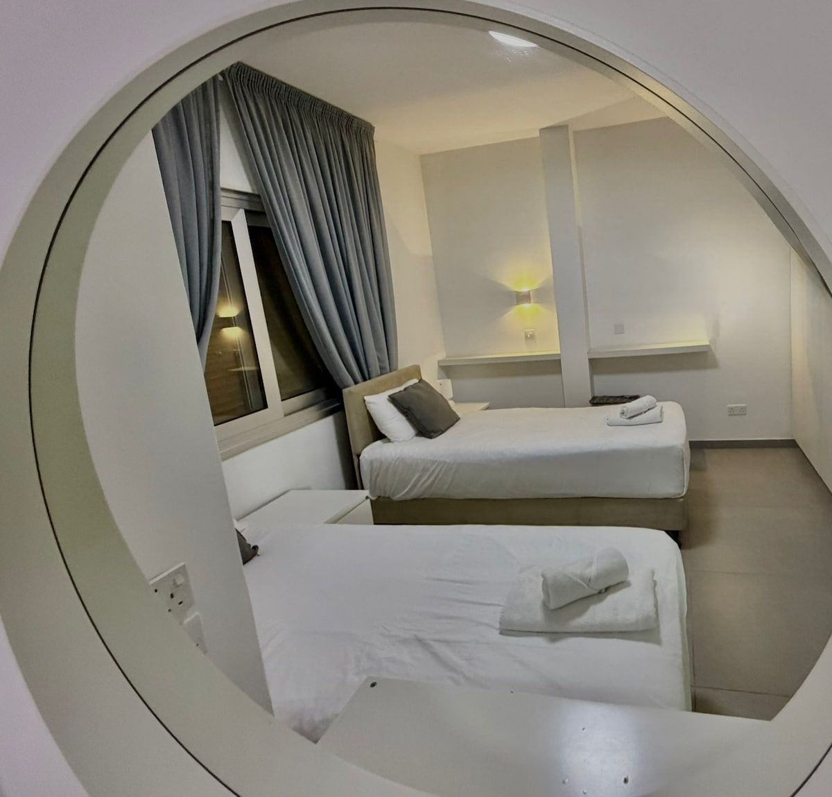 Vista circolare di una camera d'albergo con due letti, una finestra con tende, una lampada a parete e un tavolino.