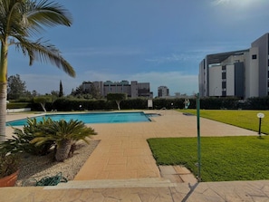 Ein Swimmingpool mit gefliestem Sonnendeck, umgeben von Grünflächen und Palmen.