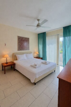 A hotel room with a bed, two bedside tables with lamps, a ceiling fan, and a view of the outdoors through a window with curtains. - Bayview Terraces – Cozy 1BR Haven (Parekklisia)