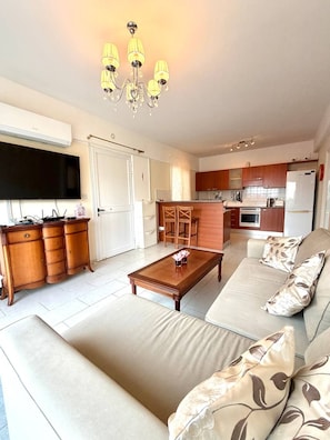 Ein Wohnzimmer mit einer Couch, einem Couchtisch und einem Fernseher. Es gibt eine Küchenzeile mit Schränken und einem Kühlschrank.