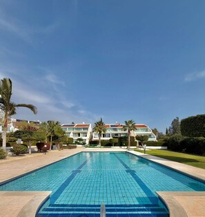 A swimming pool with a tiled deck, surrounded by palm trees and a building with multiple balconies in the background. - Bayview Terraces – Cozy 1BR Haven (Parekklisia)