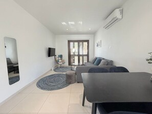 A modern living room with a sofa, a dining table, and a TV mounted on the wall. - Seaside Comfort Stay Flat Constantinos 113 (Pafos)