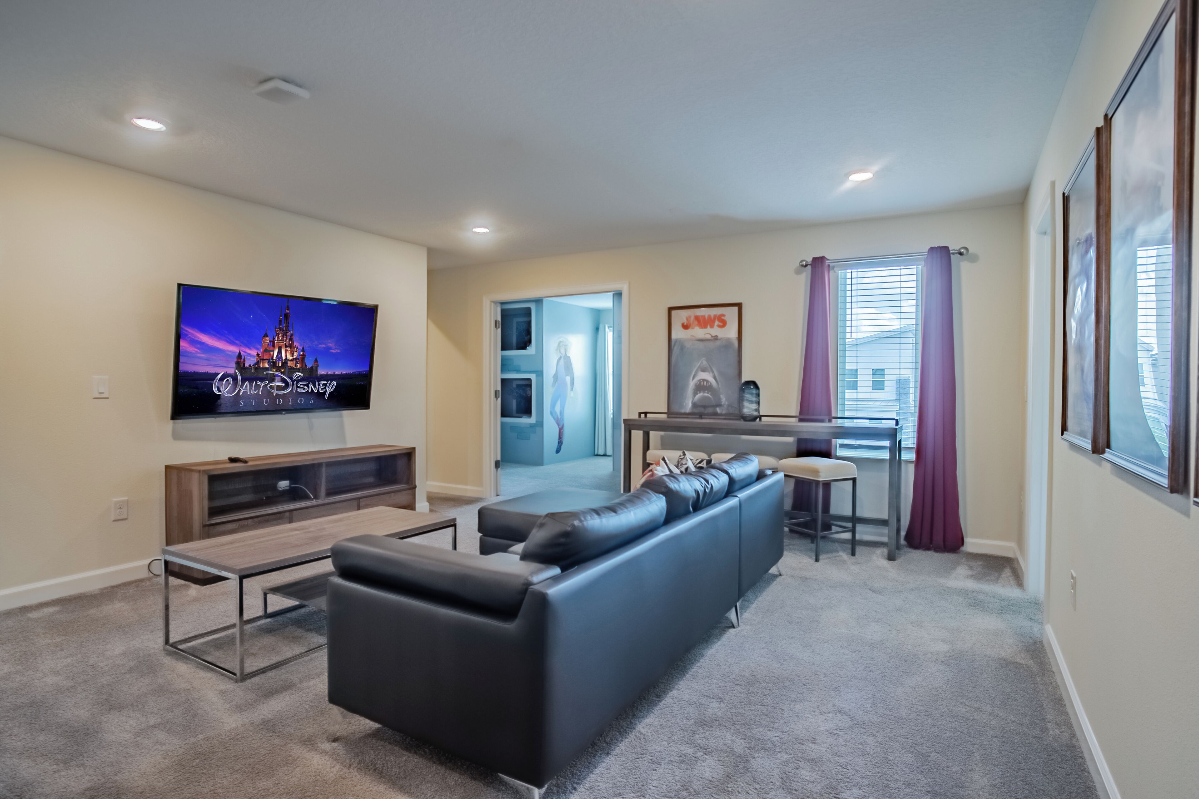A modern living room with a flat-screen TV, a black leather sofa, a wooden entertainment unit, and a dining area with a small table and chairs.