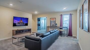 A modern living room with a flat-screen TV, a black leather sofa, a wooden entertainment unit, and a dining area with a small table and chairs.