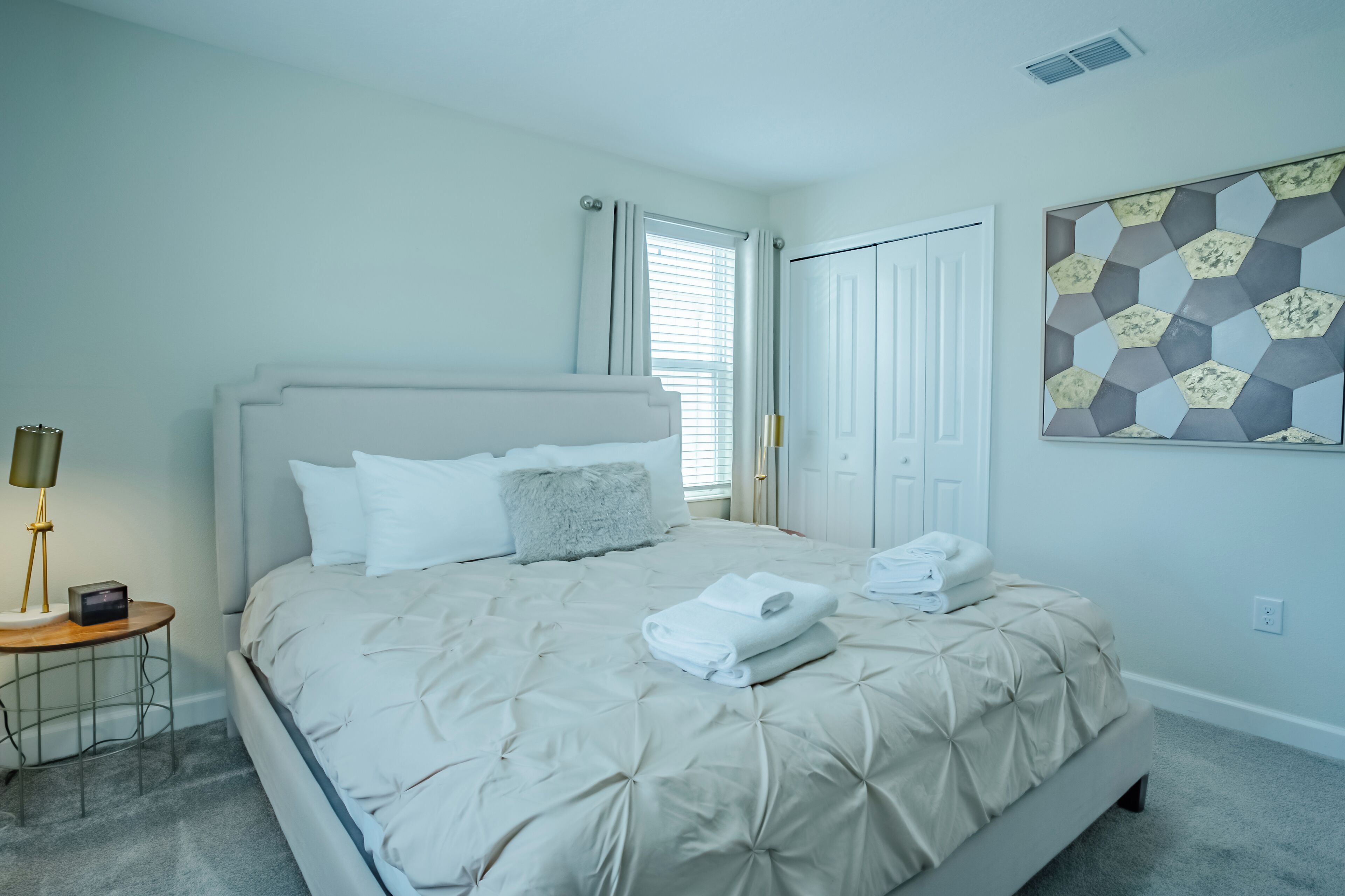 A neatly made bed with a beige comforter and pillows, a bedside table with a lamp, and a geometric wall art piece.