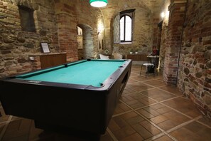 Ein Billardtisch in einem Raum mit Steinwänden, einem Fenster und einem kleinen Tisch mit Stühlen.
