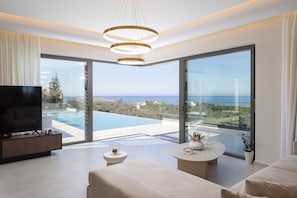 Un salón moderno con televisor, asientos, una mesita y ventanas amplias con vista al mar.