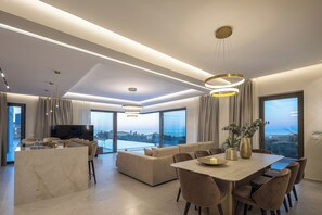 A modern living room with a dining area, large windows, and a flat-screen TV. - Simplicity of Single-Level Sea Views by etouri (Rethymno)