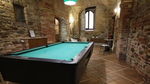Una mesa de billar en una habitación con paredes de piedra, una ventana y una pequeña mesa con sillas.