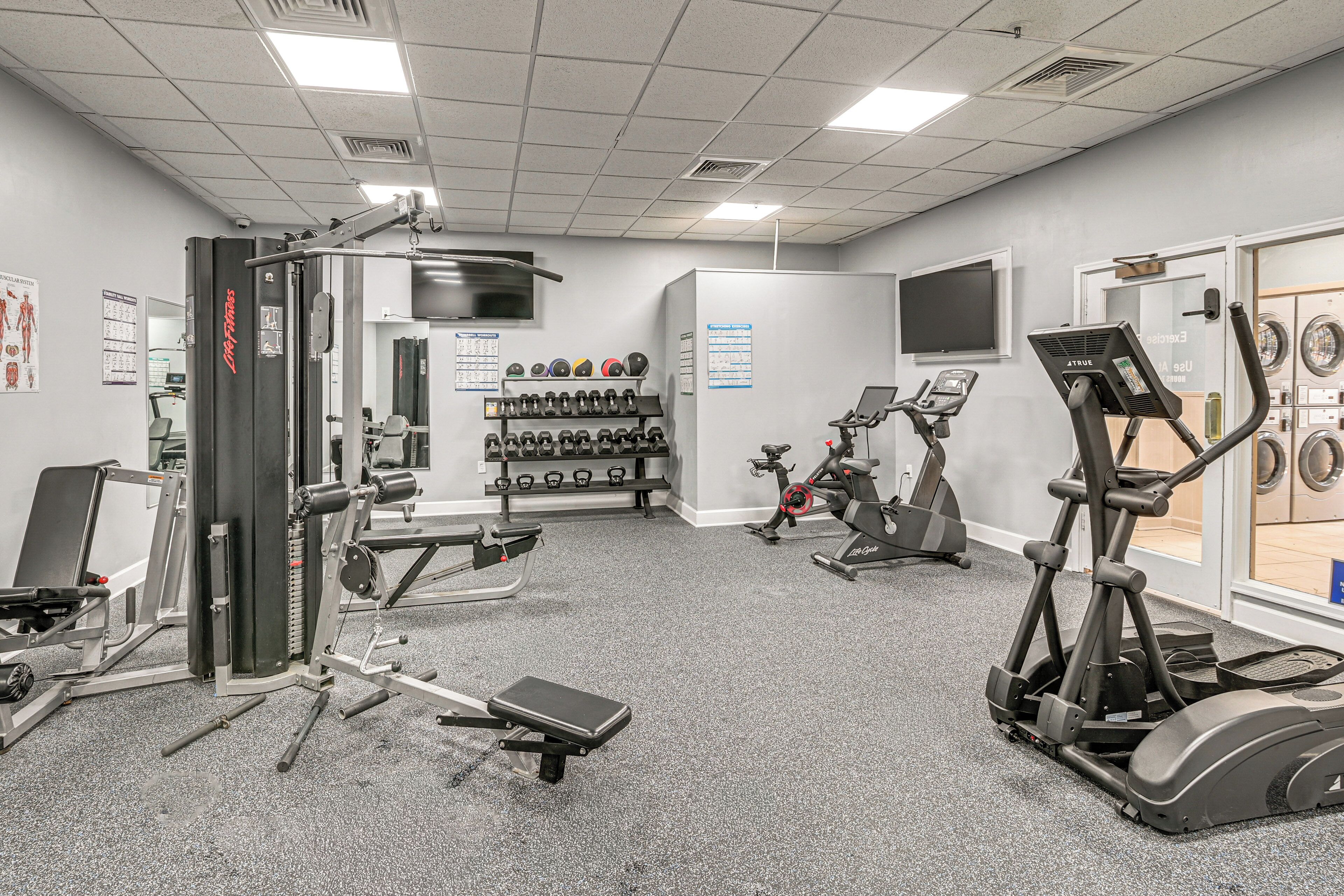 Ein Fitnessraum mit Trainingsgeräten, einem an der Wand montierten Fernseher und einer Tür zu einem Waschbereich.
